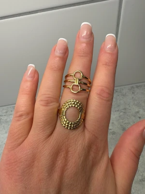 Två guldiga statementringar - Säljer två unika guldiga ringar. Den ena har en rund, strukturerad design med små nitar och öppen mitt. Den andra ringen har tre band med en dekorativ cirkeldetalj framtill. Båda ringarna är i metall och har en modern, trendig look. Rostfritt, justerbara ringar. 