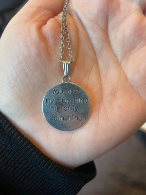 Silverfärgat halsband med solberlock - Säljer ett silverfärgat halsband med en rund berlock. Ena sidan har texten 'You are my Sunshine my only Sunshine' ingraverad och andra sidan har en sol med ett leende ansikte. Kedjan är tunn och stilren. Perfekt för dig som gillar personliga smycken.