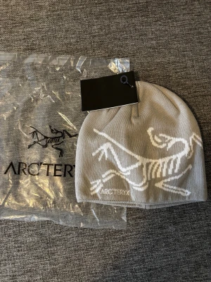 Ljusgrå mössa från Arc'teryx - Snygg ljusgrå mössa från Arc'teryx med vitt skelettmönster och logga på framsidan. Tillverkad i mjukt, stickat material som sitter skönt på huvudet. Perfekt för dig som gillar stilrena accessoarer med outdoor-vibe.