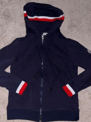Marinblå Moncler hoodie med ränder - Snygg marinblå hoodie från Moncler med dragkedja, huva och vita samt röda ränder på huvan och muddarna. Klassisk Moncler-logga på ärmen. Tillverkad i mjukt bomullsmaterial och har en avslappnad passform. Perfekt för en casual och trendig look.