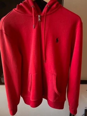 Röd hoodie från Ralph Lauren - Snygg röd hoodie från Ralph Lauren med dragkedja framtill och klassisk svart logga broderad på bröstet. Hoodien har huva med dragsko och två fickor framtill. Perfekt för en avslappnad och stilren look.