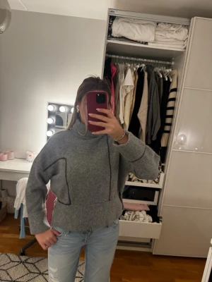 Grå stickad polotröja - Säljer en grå stickad polotröja med breda ärmar och markerade sömmar. Tröjan har en hög krage och avslappnad passform, perfekt för lager på lager. Materialet känns mjukt och mysigt, troligen ull- eller ullmix.