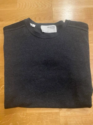 Mörkgrå tröja från Selected Homme - En stilren mörkgrå tröja från Selected Homme med rund halsringning och långa ärmar. Tröjan har ribbade muddar vid ärmslut och nederkant. Perfekt för dig som gillar en enkel och klassisk look.