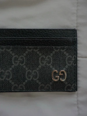 GG | Cardholder - Säljer ett svart kortfodral från med det klassiska GG-mönstret i grått och en metallogo i hörnet. Fodralet har flera kortfack och är gjort i tåligt läder med canvasdetaljer. Perfekt för dig som vill ha en stilren och lyxig accessoar. Kommer i originalförpackning, knappast använd! 