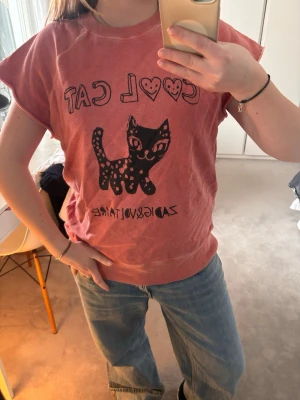Rosa t-shirt med katt från Zadig & Voltaire - Unik rosa t-shirt från Zadig & Voltaire med svart tryck av en katt och texten 'COOL CAT' med hjärtan. T-shirten har korta ärmar och avslappnad passform. Perfekt för dig som gillar statement-plagg och coola prints.