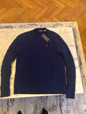 Mörkblå kabelstickad tröja Polo - Mörkblå kabelstickad tröja från Polo Ralph Lauren med rund halsringning och ribbade muddar. Klassisk röd logga broderad på bröstet. Perfekt för lager på lager och riktigt snygg till jeans eller chinos.