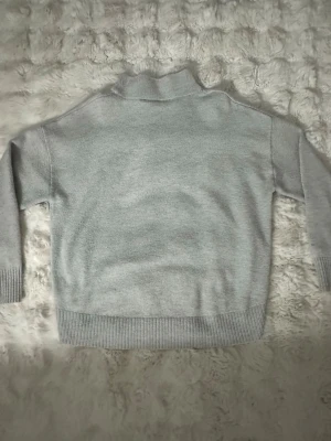 Ljusgrå stickad polotröja - Mysig ljusgrå stickad polotröja med avslappnad passform. Tröjan har ribbade muddar vid ärmslut och nederkant samt en hög krage. Perfekt för kalla dagar och enkel att matcha med olika outfits.