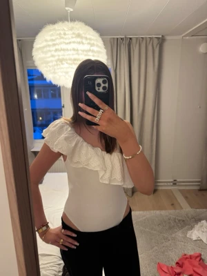 Vit off shoulder body med volang - Säljer en vit body med en axel och stor volangdetalj. Bodyn har broderade hålmönster på volangen och är i stretchigt material. Perfekt för dig som vill ha något trendigt och bekvämt med unik design.