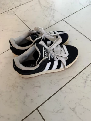 Svarta Adidas sneakers med vita ränder - Klassiska svarta Adidas sneakers med vita ränder och vit sula. Använda ett fåtal gånger och är i bra skick! Lite defekter i självaste skon utöver det är det som nya 