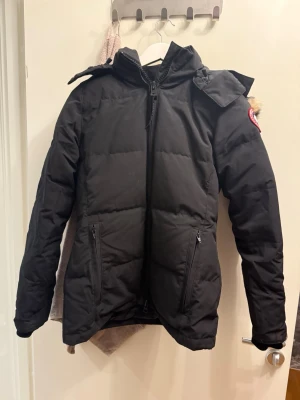 Canada Goose - Säljer en svart dunjacka från Canada Goose med huva och avtagbar fuskpäls. Jackan har dragkedja framtill, två fickor med dragkedja och klassisk patch på ärmen. Perfekt för kalla vinterdagar och riktigt snygg passform.