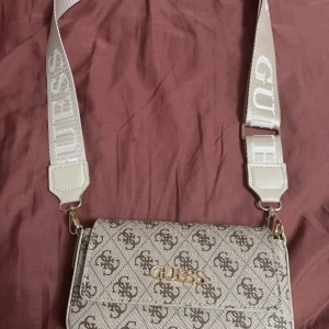 Beige axelväska  - Snygg beige axelväska från Guess med bred axelrem där märkesnamnet står i vitt. Väskan har ett klassiskt logomönster i brunt och guldigt Guess-emblem framtill. Perfekt storlek för det viktigaste och ger en trendig touch till din outfit. Pris kan diskuteras.