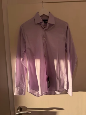 Ljuslila skjorta från Dressmann - Klassisk ljuslila skjorta från Dressmann med krage och knäppning framtill. Skjortan har lång ärm och är tillverkad i bomull för en bekväm känsla. Perfekt för en clean och stilren look.