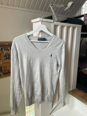 Ljusgrå v-ringad tröja Polo Ralph Lauren - Ljusgrå v-ringad tröja från Polo Ralph Lauren med den klassiska blå loggan broderad på bröstet. Tröjan har långa ärmar, ribbade muddary och är tillverkad i ett mjukt, tunt material som passar perfekt till både finare och vardagliga stunder!                                       Byten kan vara intressant!