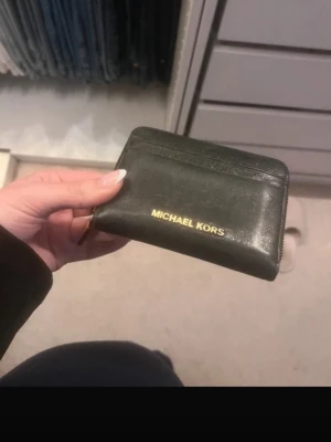 Svart plånbok från Michael Kors - Snygg svart plånbok från Michael Kors i läder med guldfärgad logga framtill. Plånboken har dragkedja, flera fack för kort och mynt samt stilren design. Perfekt storlek för att ha med sig i fickan eller väskan. Köpt på zalando för 1095