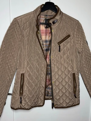 Beige quiltad jacka från Saki - Stilren beige quiltad jacka från Saki med bruna detaljer och dragkedjor. Jackan har ett rutigt foder i flera färger och tre fickor med dragkedja framtill. Perfekt för dig som gillar klassisk men trendig look.