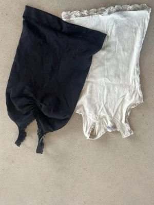 2-pack bodys med spetsdetaljer - Säljer två bodys från Lindex i storlek 134/140. Ena är svart med spetskant och rynk framtill, den andra är vit med ribbad struktur och spets runt hals och ärm. Perfekta under kläder, mjuka och stretchiga i materialet.