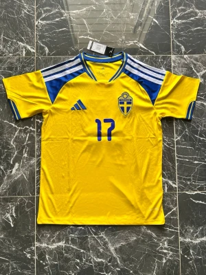 Sverige matchtröja Adidas gul/blå - Gul matchtröja från Adidas med blå detaljer och nummer 17 på bröstet. Tröjan har Sveriges landslagsemblem på vänster sida och klassiska Adidas-ränder på axlarna. Tillverkad i lätt och ventilerande material, perfekt för fotboll eller träning.
