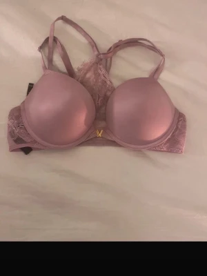 Rosa push-up bh från Victoria's Secret - Snygg rosa push-up bh från Victoria's Secret med vadderade kupor och spetsdetaljer på sidorna. Bh:n har justerbara axelband och en liten guldfärgad V-detalj framtill. 