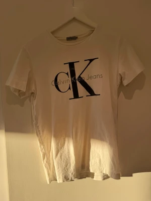 Vit Calvin Klein Jeans t-shirt - Vit t-shirt från Calvin Klein Jeans med stor svart CK-logga och text på bröstet. Klassisk rund halsringning och korta ärmar. Tillverkad i mjuk bomull som känns skön mot huden. Perfekt för en clean och stilren look.