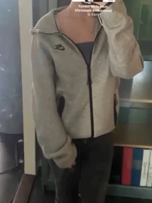 Grå Nike hoodie med dragkedja - Säljer en grå hoodie från Nike med svart dragkedja och svarta detaljer vid fickorna. Hoodien har en huva och Nike-logga på bröstet. Perfekt för en avslappnad och sportig stil. Materialet är mjukt och bekvämt, troligen bomullsblandning.