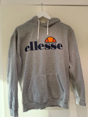 Grå Ellesse hoodie med logga - Säljer en grå hoodie från Ellesse med stor logga i mörkblått och orange framtill. Hoodien har huva med vita dragsnören och en stor magficka. Tillverkad i mjuk bomullsblandning, perfekt för chill dagar eller när du vill ha en sportig look.