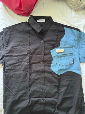 Svart skjorta med blå denimficka - Unik svart skjorta från Aelfric Eden med en stor blå denimficka och kontrasterande axelparti. Skjortan har klassisk krage, knappar framtill och korta ärmar. Perfekt för dig som gillar statement-plagg och vill sticka ut. Aldrig använd.