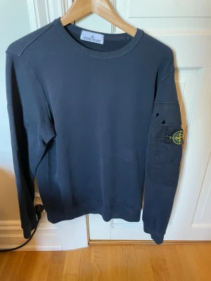 Mörkblå sweatshirt från Stone Island - En svart långärmad sweatshirt från Stone Island med rund halsringning och klassisk logotyp-patch på vänster ärm. Tröjan har en ficka på ärmen och är tillverkad i mjuk bomull för en bekväm känsla. Köpt för 3799kr på NK Classic Man i Göteborg . Digitalt kvitto finns