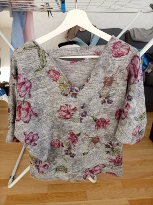 Grå blommig topp med v-ringning - Grå topp med v-ringning och trekvartsärm, dekorerad med blommigt mönster i rosa och lila toner. Mjuk och skön känsla, perfekt för en avslappnad stil. Lätt att matcha med jeans eller kjol.