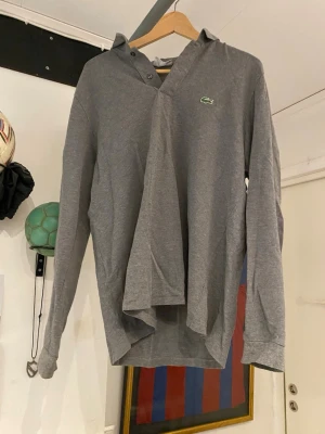 Grå henley från Lacoste - Säljer en grå långärmad henley från Lacoste med klassisk knappslå och den ikoniska gröna krokodilloggan på bröstet. Skjortan är i mjuk bomull och har en avslappnad passform som passar perfekt till vardagslooken.