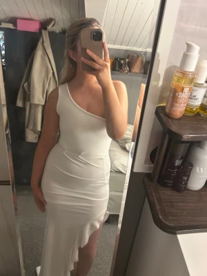 Vit långklänning med slits - Elegant vit långklänning med asymmetrisk axel och hög slits framtill. Klänningen har en figurnära passform och volangdetalj längs slitsen. Perfekt för dig som vill sticka ut med en stilren och modern look.