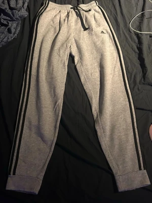 Gråa Adidas mjukisbyxor med svarta ränder - Gråa mjukisbyxor från Adidas med klassiska svarta ränder längs sidorna och Adidas-logga på vänster lår. Byxorna har resår och dragsko i midjan för skön passform. Perfekta för chill eller träning 