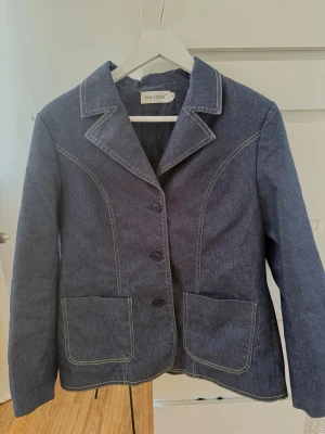 Mörkblå jeansjacka från Prestige - Snygg mörkblå jeansjacka från Prestige med klassisk krage, tre knappar framtill och två stora fickor. Jackan har vita kontrastsömmar och figurnära passform som ger en stilren look. Materialet har glitter i sig. 