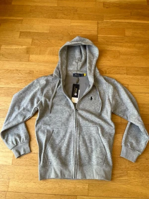 Grå zip-hoodie från Polo Ralph Lauren - Snygg grå hoodie med dragkedja från Polo Ralph Lauren. Klassisk design med huva och dragsnören, två fickor fram och broderad logga på bröstet. Mjuk bomullsblandning som är skön att ha på sig. Perfekt för en avslappnad och stilren look. Pris kan diskuteras. Vid frågor eller funderingar är det bara att höra av sig. 