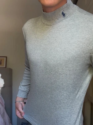 Ljusgrå polotröja från Ralph Lauren - Snygg ljusgrå polotröja från Ralph Lauren med diskret logga broderad vid kragen. Tröjan är långärmad och har en smal passform. Tillverkad i mjukt och stretchigt material som sitter skönt på kroppen. (Passform: jag på bilden är 190 lång och väger 80kg)