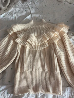 Krämvit stickad tröja med volang - Supersöt krämvit stickad tröja med breda volanger över axlar och bröst. Tröjan har ribbstickad struktur, hög hals och långa ärmar. Perfekt för dig som gillar romantisk och trendig stil. Materialet känns mjukt och bekvämt.