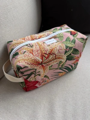 Tropisk necessär från Emelo - Necessär i tyg med färgglatt blommönster i rosa, grönt, gult och rött. Rektangulär form med vit dragkedja och praktiskt handtag i beige. Perfekt för smink eller småprylar. Märke: Emelo. Innerfack finns. Mått ca 27x17x12cm