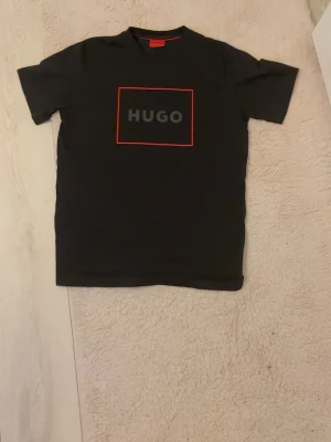 Hugo boss Tshirt - Svart t-shirt från HUGO med en stilren röd ram och HUGO-logga på bröstet. Klassisk passform och rund halsringning. Perfekt för en trendig och enkel look. Storlek L passar M