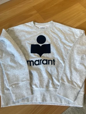 Grå sweatshirt från Marant Étoile - Snygg grå sweatshirt från Marant Étoile med stort svart logotryck på bröstet. Tröjan har rund halsringning, ribbade muddar och är tillverkad i mjukt bomullsmaterial. Perfekt för en avslappnad och trendig look.