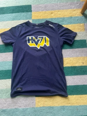 Blå HV71 t-shirt från Craft - Mörkblå t-shirt från Craft med HV71-logga i gult och vitt på bröstet. Klassisk rund hals och korta ärmar. Perfekt för dig som vill visa vilket lag du hejar på. Tillverkad i mjuk bomull för skön känsla.