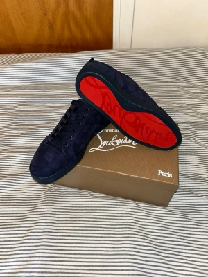 Mörkblå Louboutin sneakers i mocka - Snygga mörkblå sneakers från Christian Louboutin i mjuk mocka med klassisk röd sula och svarta skosnören. Låg modell med diskret logga på insidan och stilren design. Perfekta för dig som vill ha något exklusivt och unikt i garderoben.