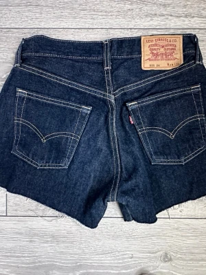 Levi's mörkblå denim shorts - Säljer ett par klassiska blå denimshorts från Levi's med tydliga kontrastsömmar och den ikoniska läderlappen bak i midjan. Shortsen har normal passform och är tillverkade i slitstark bomull. Perfekta för sommaren och ger en tidlös look. Strl 29” men små, se bild på midjeband. 