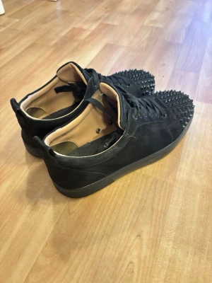 Louboutin  - Säljer ett par svarta sneakers i mocka med coola svarta nitar över tån. Skorna har snörning och en platt sula. Insidan är ljusbeige och de har en modern, edgy look som sticker ut.
