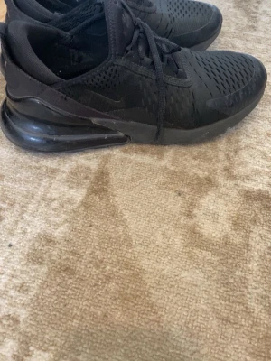 Svarta Nike Air Max 270 sneakers - Säljer ett par helsvarta Nike Air Max 270 sneakers med stor synlig Air-enhet i hälen. Skorna har mesh-ovandel för bra ventilation, klassisk Nike-logga på sidan och snörning. Snygg och sportig design som passar till streetwear.