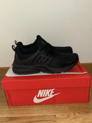 Svarta Presto sneakers - Snygga helsvart Presto med mesh-ovandel och plastdetaljer på sidorna. Skorna har en låg silhuett, rund tå och klassisk Nike-logga på både plös och sidor. Perfekta för dig som gillar stilrena och bekväma sneakers.
