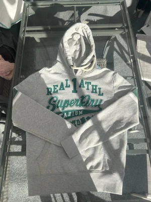 Superdry Hoodie Grå/Grön – Y2K Streetwear Strl L - Säljer en Superdry hoodie i mycket bra skick. Grym hoodie för sommarkvällar och passar perfekt till en clean streetwear outfit. Den grå färgen med gröna vintage-inspirerade tryck ger en riktigt snygg y2k vibe och gör att den sticker ut lagom mycket.  Mjuk och bekväm med fleecefodrad insida, kangaroo pocket och oversized känsla beroende på hur man vill bära den. Enkel att matcha med jeans, cargos eller shorts för en avslappnad casual look. Perfekt för dig som gillar retro, streetstyle och trendig