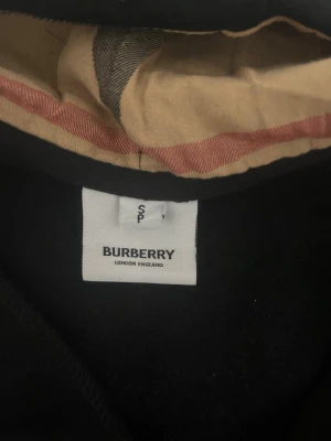 Svart hoodie från Burberry med dragkedja - Stilren svart hoodie från Burberry med dragkedja och klassiskt rutigt foder i huvan. Hoodien har dragsko vid huvan och ribbade muddar. Perfekt för dig som vill ha en lyxig och tidlös look. Märkeslapp kvar på plagget.