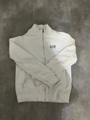 Vit zip hoodie från EA7 Emporio Armani - Snygg vit hoodie med dragkedja från EA7 Emporio Armani. Modellen har hög krage, två fickor framtill och diskret EA7-logga på bröstet. Perfekt för en clean och stilren look. Tillverkad i mjukt material för extra komfort.