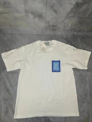Vit t-shirt från Casablanca - Vit t-shirt från Casablanca med en blå grafisk ruta på bröstet. Klassisk passform med rund hals och korta ärmar. Perfekt för dig som gillar stilrena plagg med en twist. Materialet känns mjukt och skönt mot huden.