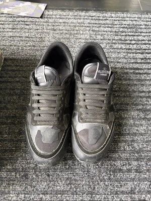 Valentino Garavani svarta sneakers - Snygga svarta sneakers från Valentino Garavani med camomönster och detaljer i mocka och läder. Skorna har en chunky sula och klassisk låg profil med snörning. Perfekta för dig som vill ha en exklusiv och streetinspirerad look.