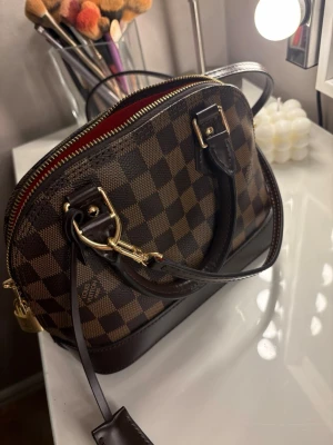 Brun handväska - Snygg handväska klassiskt brunt Damier-mönster. Väskan har rundad form, mörkbruna läderdetaljer, guldfärgade metalldetaljer och röd textilinsida. Kommer med både handtag och axelrem för flexibel användning. Helt oanvänd